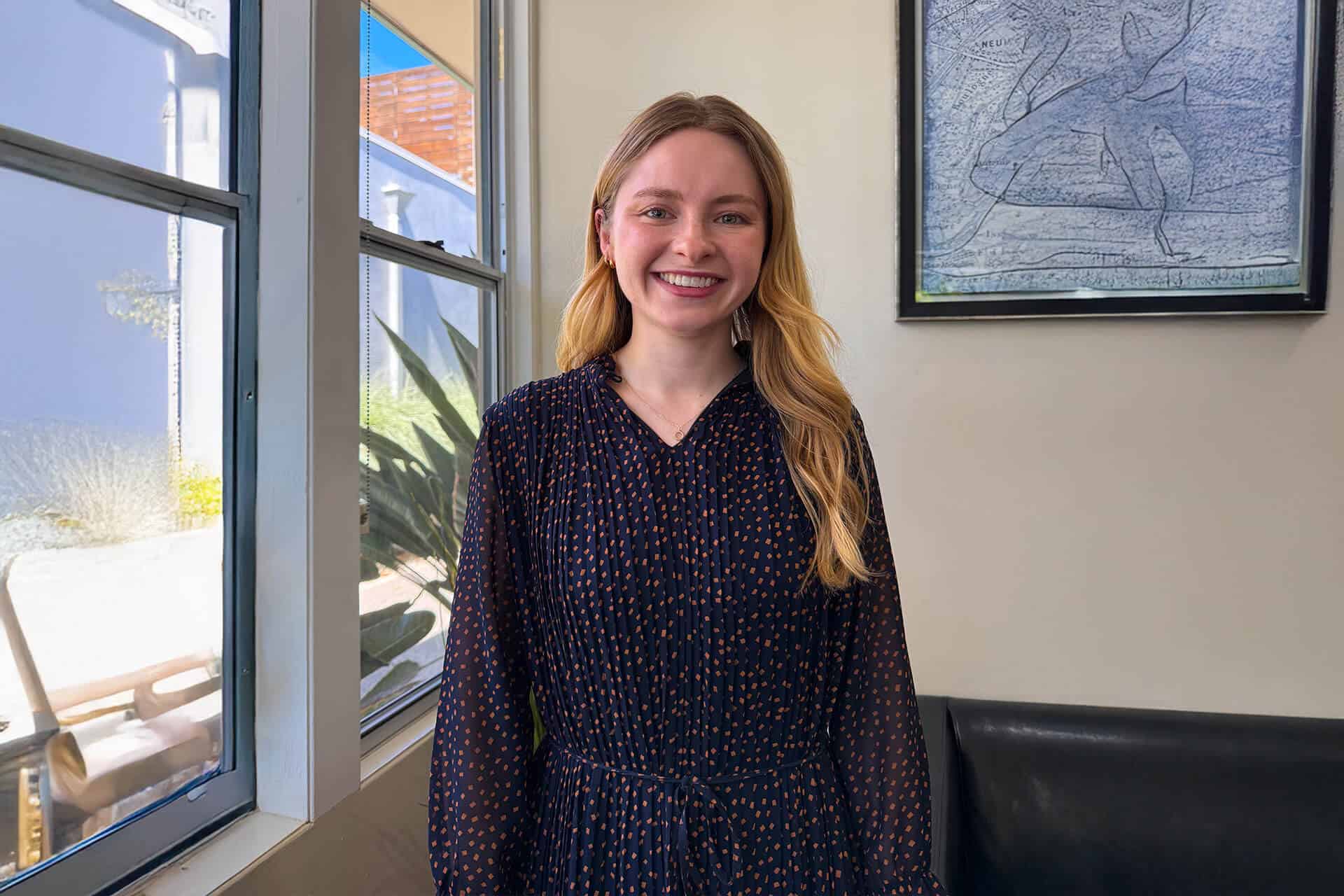 Bright smiling woman indoors near window, casual professional attire, positive expression, natural light, modern office environment, friendly presentation, confident demeanor.